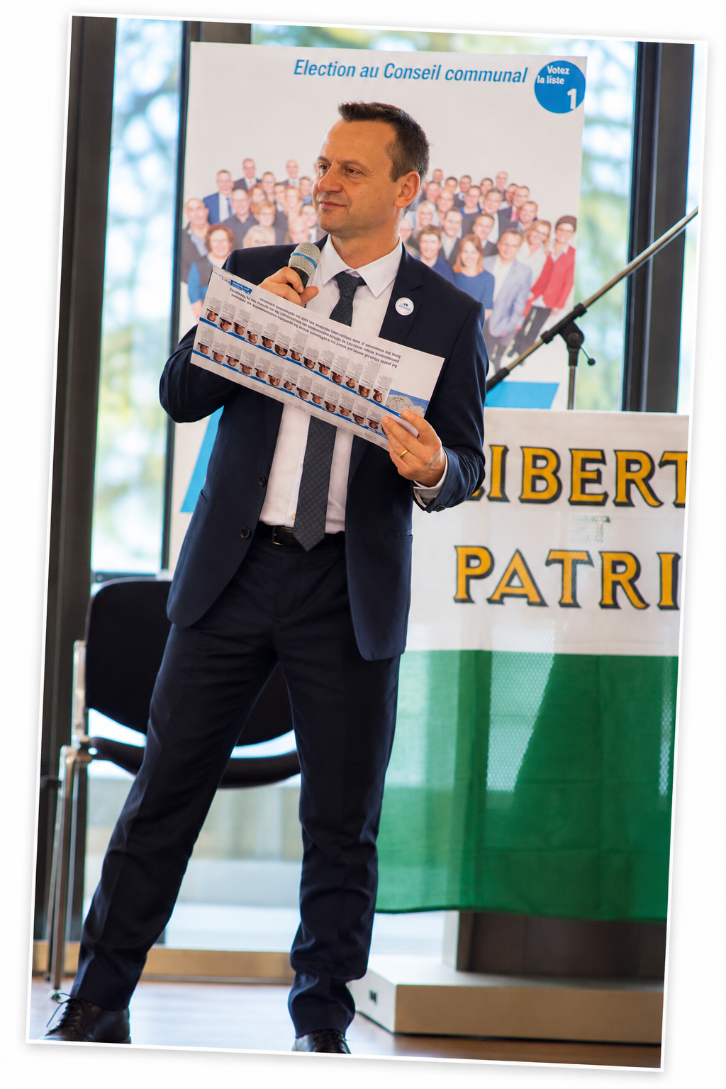 Yves Depallens speaking at a political event in Montreux, holding a microphone in front of a poster about the Communal Council elections.
